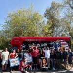A group of people, with protest signs, standing in front of a red bus that reads "SAVING THE MOTDERF**KING WORLD!" and "MAGABUS."